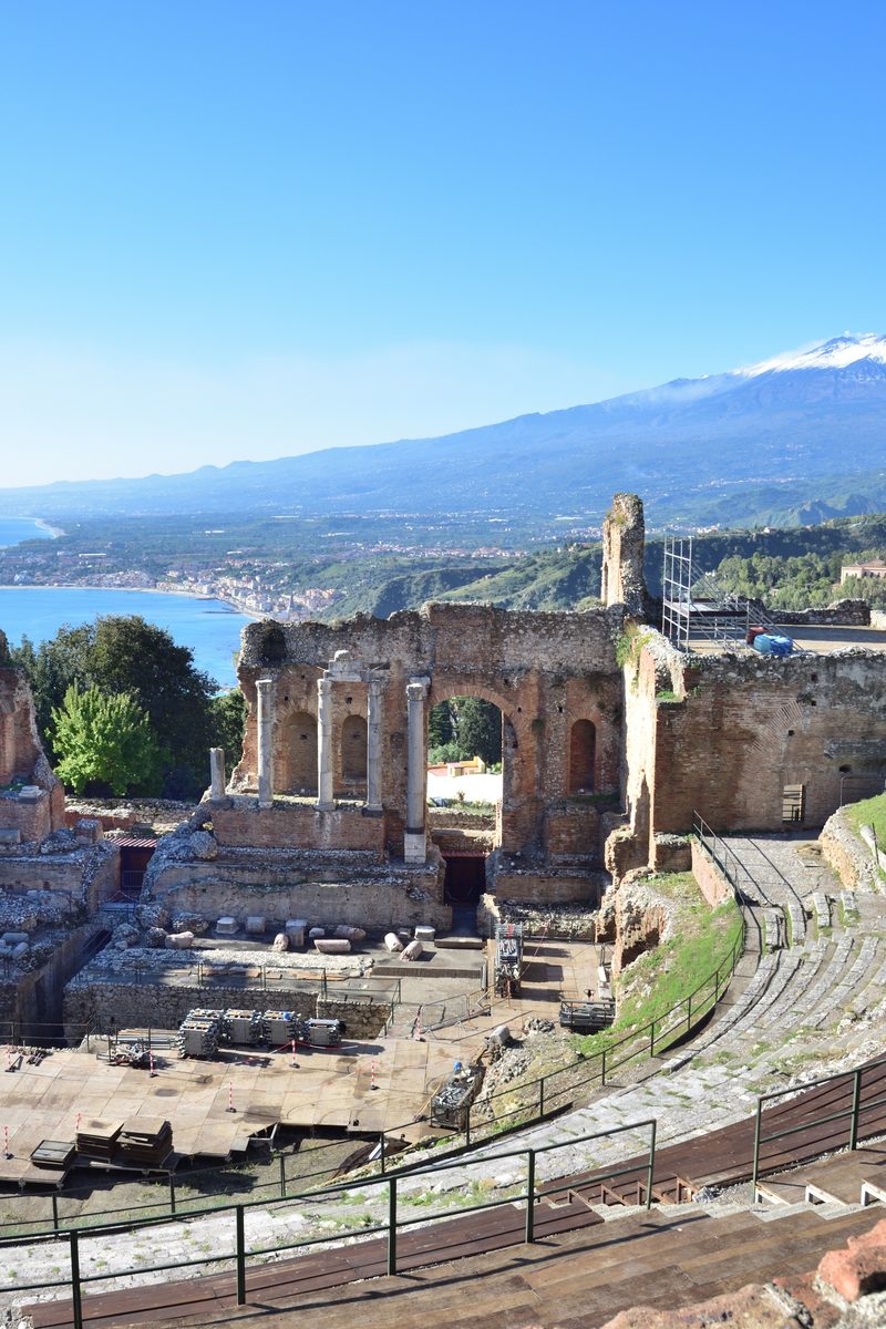 Les gradins taillés dans la roche du Théâtre Antique de Taormine, avec le mont Etna et la mer Ionienne encadrés au-delà du mur de scène en ruine